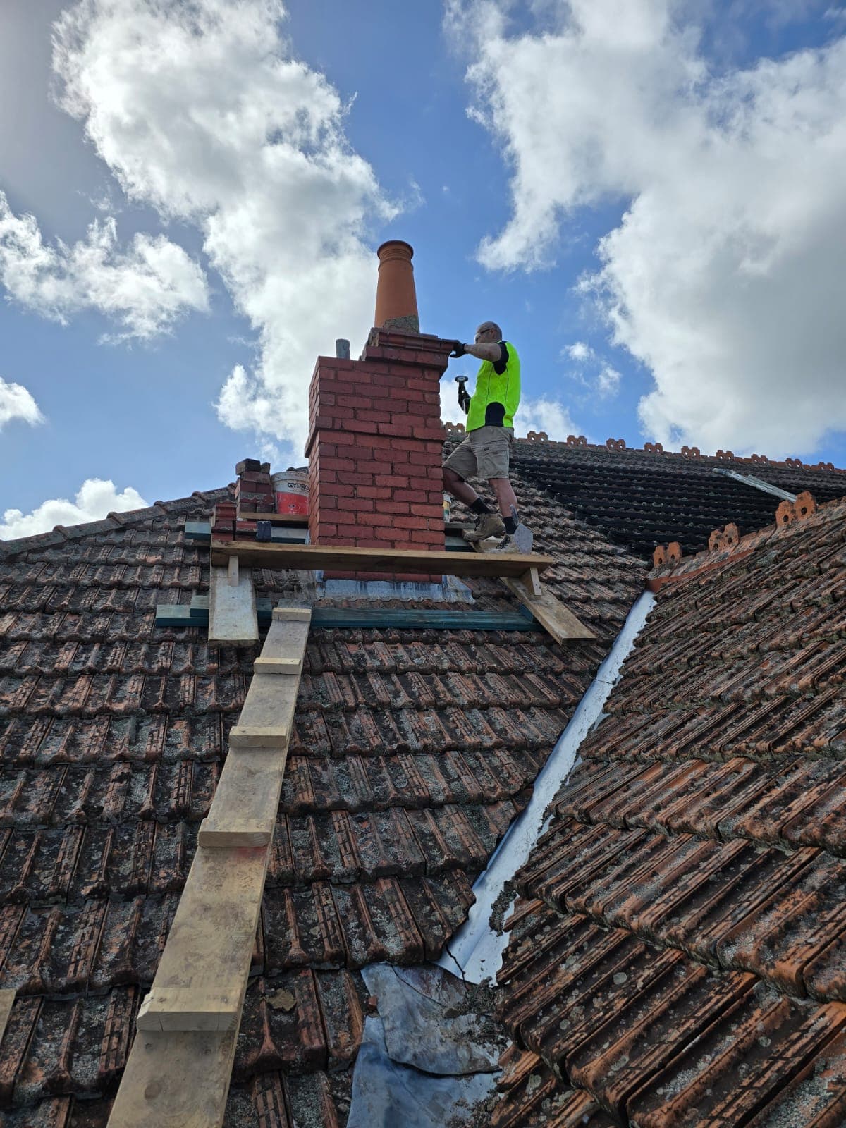 Bricklayer rebuilding a tall red-brick heritage chimney on a tiled Melbourne roof by Goodwin Bricklaying